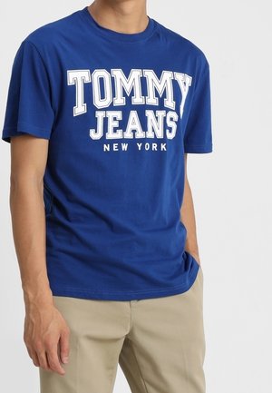 Mann trägt ein blaues Rundhals-T-Shirt mit "TOMMY JEANS NEW YORK" in Weiß aufgedruckt und beige Hose, Hand in der Tasche.