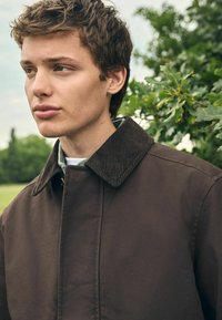 Chaqueta marrón con cuello de pana, que presenta una textura suave y un cierre frontal de cremallera. El modelo está de pie frente a un fondo de vegetación, iluminado de forma natural.