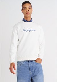 Sudadera blanca con cuello redondo y logo bordado de "Pepe Jeans" en azul. Combinada con una camisa de cuadros azul debajo y jeans de un azul claro.
