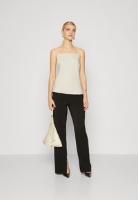 Cream-colored spaghetti strap top, black high-waisted trousers, and pointed black heels. Model holds a cream bag. Simple, elegant design.