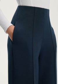 Morske hlače z visokim pasom z gladko teksturo, ki imajo stranske žepke in ostre plisirane detajle, v kombinaciji s prilegajočo belo majico z dolgimi rokavi.