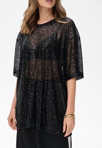 Kobieta w czarnej, przezroczystej bluzce z cekinami nosząca czarny biustonosz, połączona z czarnymi spodniami oraz trzema złotymi bransoletkami na prawej ręce.