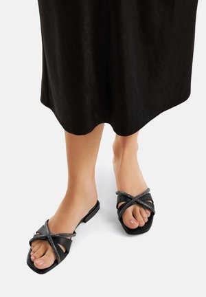 Crne ravne sandale z zasnovano na prepletenih trakovih z metalnimi detajli. Traki so narejeni iz gladkega materiala, podplat pa je kvadratne oblike.