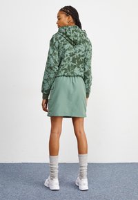 Zöld, batikolt kapucnis pulóver kenguruzsebbel, ami egy jól illeszkedő zöld szoknyával és bordázott szürke zoknikkal van párosítva, fehér sneakers-szel.