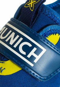 Scarpa sportiva blu con tomaia in tessuto testurizzato, ampia cinghia blu con il logo bianco "MUNICH" e dettagli in giallo.