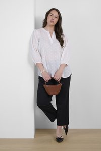 Chemisier blanc transparent avec des pois texturés, col montant et manches bouffantes, associé à un pantalon noir à jambes larges et des ballerines noires pointues. Sac à main marron.