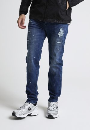 Uomo che indossa jeans blu consumati, sneakers New Balance bianche e grigie, e una giacca nera con le mani nelle tasche.