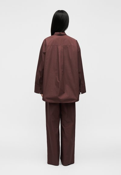 Personne aux longs cheveux noirs, vue de dos, portant une chemise marron foncé ample et un pantalon large assorti sur un fond uni.