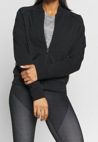 Musta vetoketjullinen takki pehmeästä kankaasta, jossa on pyöreä kaulus, yhdistettynä teksturoituihin mustiin legginsseihin, joissa on geometrinen kuvio.