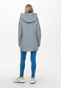 Abrigo gris con capucha, de corte entallado, mangas largas y dobladillo recto. Lleva puesto sobre jeans de mezclilla azul y zapatillas de color claro.