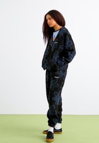 Completo in pile nero con grandi motivi floreali blu; giacca e pantaloni a vestibilità comoda, abbinati a scarpe nere e calze bianche.