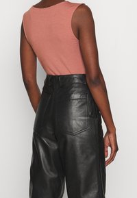 Rust-colored sleeveless top paired with high-waisted black leather trousers, featuring back pockets and a smooth, shiny texture.
