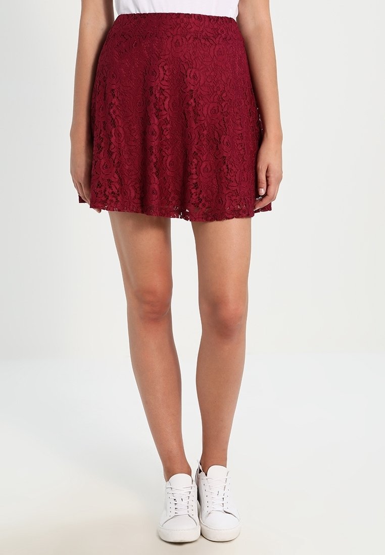 Femme portant une jupe en dentelle rouge à motifs floraux et des baskets blanches, debout devant un fond clair uni.