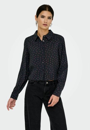 Vrouw in een zwarte blouse met kleine roze strikpatronen en zwarte hoge taille jeans, staand tegen een effen achtergrond.