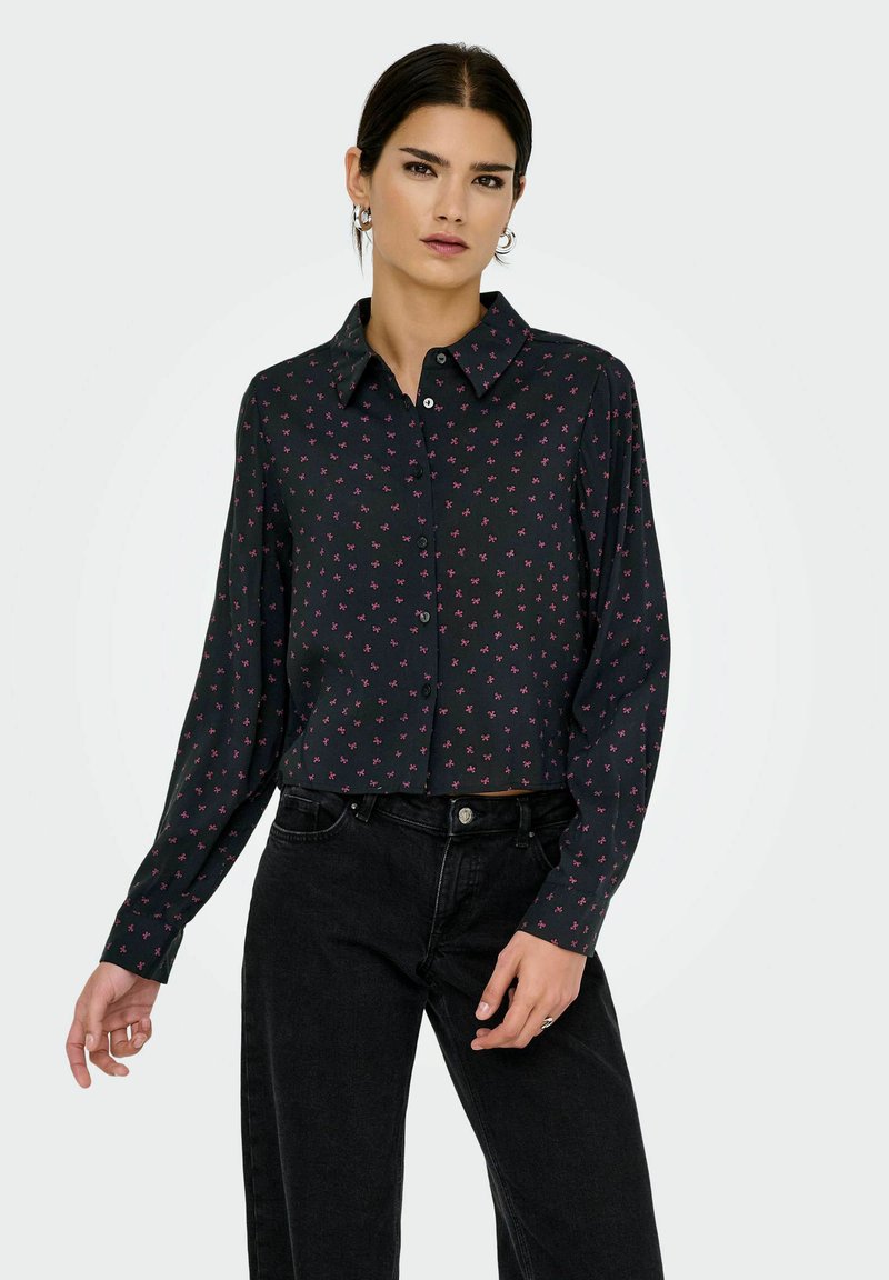 Femme portant une chemise noire à boutons avec de petits motifs de nœuds roses et un jean noir taille haute, se tenant devant un fond uni.