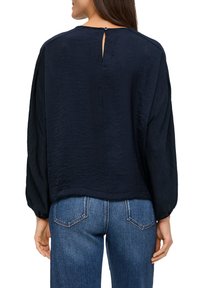 Navybluse mit strukturierter Oberfläche, lockerer Passform, Keyhole-Detail am Rücken mit einem Knopf und langen Ärmeln, kombiniert mit blauen Jeans.