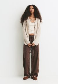 Femme avec de longs cheveux bouclés portant un cardigan beige à boutons, un débardeur blanc, un pantalon large marron et des chaussures à lacets marron, se tenant contre un mur blanc.