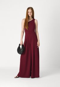 Une combinaison sans manches bordeaux avec un décolleté asymétrique, des jambes larges et des plis. Associée à un sac à main noir texturé.