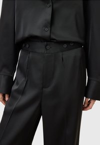 Blusa e pantaloni in seta nera, caratterizzati da una texture liscia, dettagli con bottoni in vita e una vestibilità sagomata con pieghe laterali.