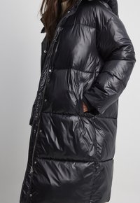 Schwarze Daunenjacke mit glänzender Oberfläche, ausgestattet mit einer Kapuze, großen gesteppten Abschnitten, seitlichen Taschen und einem Reißverschluss auf der Vorderseite.