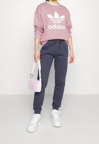 Różowa bluza z białym logo Adidas; granatowe spodnie dresowe z sznurkiem; jasnoróżowa torebka; białe sneakersy z teksturowaną podeszwą.