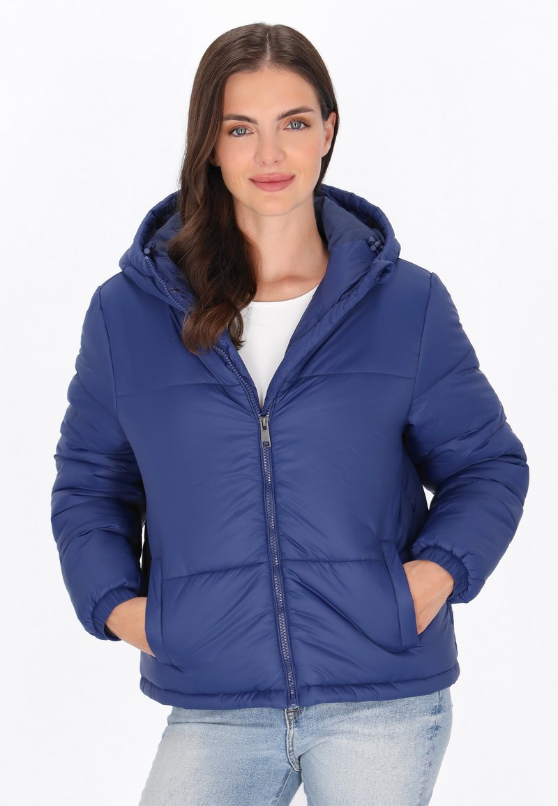 Femme portant une veste matelassée à capuche bleue avec des poches devant, une chemise blanche et un jean bleu clair, debout les mains dans les poches.