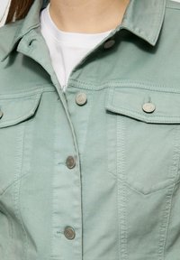 Helle grüne Jeansjacke mit silbernen Druckknöpfen, Brusttaschen und einem strukturierten Kragen. Glatte Textur mit detaillierten Nähten.