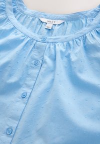 Hellblaue Bluse zum Knöpfen aus weichem Stoff; verfügt über einen runden Ausschnitt, eine dezente Textur und kleine Knopfdetails entlang der Vorderseite.