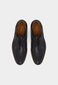 Μαύρα δερμάτινα παπούτσια brogue με πουά πτερύγια και καφέ εσωτερική επένδυση, απεικονιζόμενα από πάνω σε λευκό φόντο.