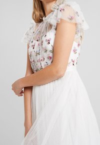 Valkoinen mekko, jossa kirjailtuja kukka-aiheisia yksityiskohtia, läpikuultavat pilkkukuvioidut hihat ja laskostettu hame. Pinta on teksturoitu, ja olkapäillä on pehmeitä röyhelöitä.