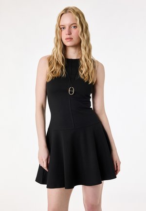 Jeune femme aux longs cheveux blonds ondulés, portant une robe noire sans manches et un pendentif, posant devant un fond blanc uni.