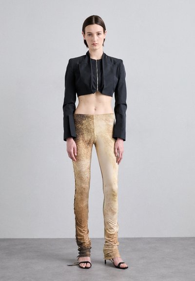 Black cropped jacket with front zip, paired with form-fitting beige pants featuring a textured, gradient earth-tone pattern and gathered detailing at the ankles.