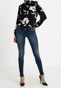Una donna sta in piedi con una mano in tasca, indossando una camicia nera a maniche lunghe con motivo floreale, jeans skinny blu scuro e scarpe con tacco alte nere.