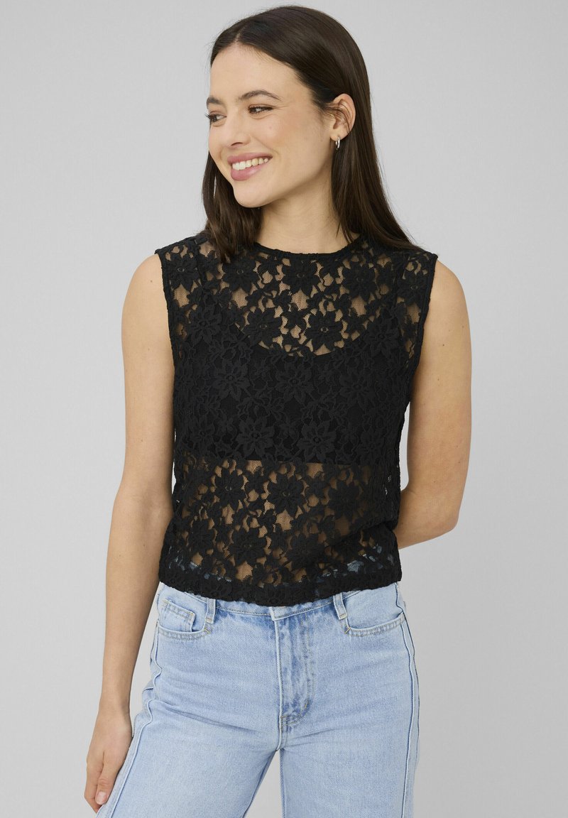 Jeune femme souriante, portant un haut sans manches en dentelle florale noire et un jean taille haute bleu clair, sur un fond gris uni.