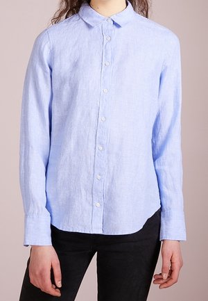 Jasnoniebieska koszula z długim rękawem, zapinana na guziki, z kołnierzykiem, noszona przez osobę stojącą na jednolitym tle.