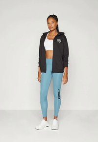 Γυναίκα που στέκεται, φορώντας μαύρο φούτερ με φερμουάρ Puma, άσπρο αθλητικό σουτιέν, μπλε κολάν Puma και άσπρα αθλητικά παπούτσια, μπροστά από έναν απλό φόντο.