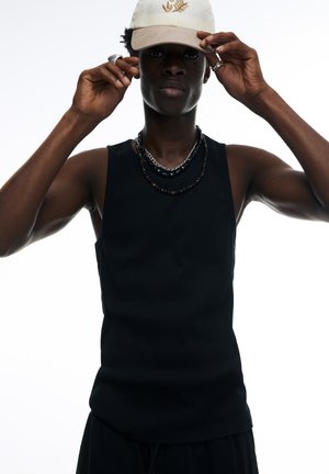 Débardeur noir sans manches avec une texture lisse, design ajusté. Porté avec des colliers superposés et une casquette crème arborant un logo discret.