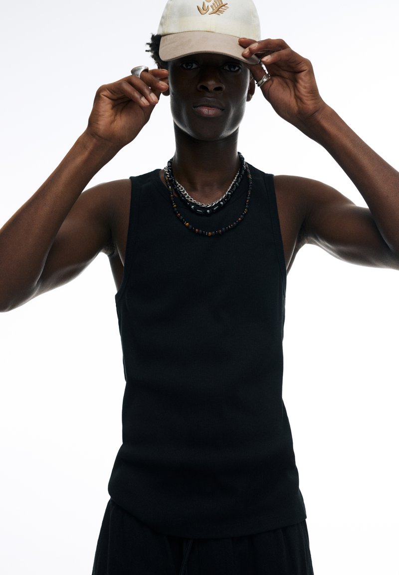 Black sleeveless tank top with a smooth texture, fitted design. Worn with layered necklaces and a cream cap featuring a subtle logo.