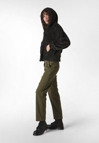 Giacca nera pelosa con cappuccio, pantaloni verdi e stivali neri stringati. La giacca presenta una vestibilità rilassata e tasche laterali. Texture del tessuto morbida.