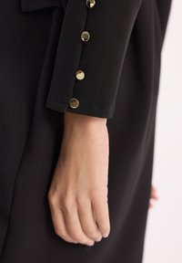 Czarny rękaw odzieży o gładkiej fakturze. Złote okrągłe guziki zdobią mankiet, dodając wyrazistego akcentu do projektu. Ręka częściowo widoczna.