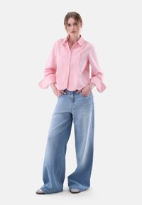 Pink collared button-up shirt with long sleeves, paired with wide-leg light blue denim jeans. Model poses with hands in pockets.