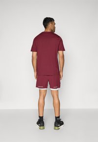 T-shirt à manches courtes bordeaux et shorts assortis. Tissu lisse, coupe décontractée, avec des accents blancs au bas des shorts et du texte de la marque. Baskets noires.