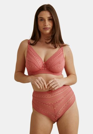 Femme portant un ensemble assorti de lingerie rose en dentelle avec un soutien-gorge et un slip taille haute, debout, les mains doucement jointes à la taille, sur un fond uni.