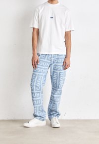Camiseta de algodón blanca con un logotipo azul, combinada con pantalones de mezclilla azul claro con un atrevido estampado repetido de "HUGO". Zapatillas blancas.