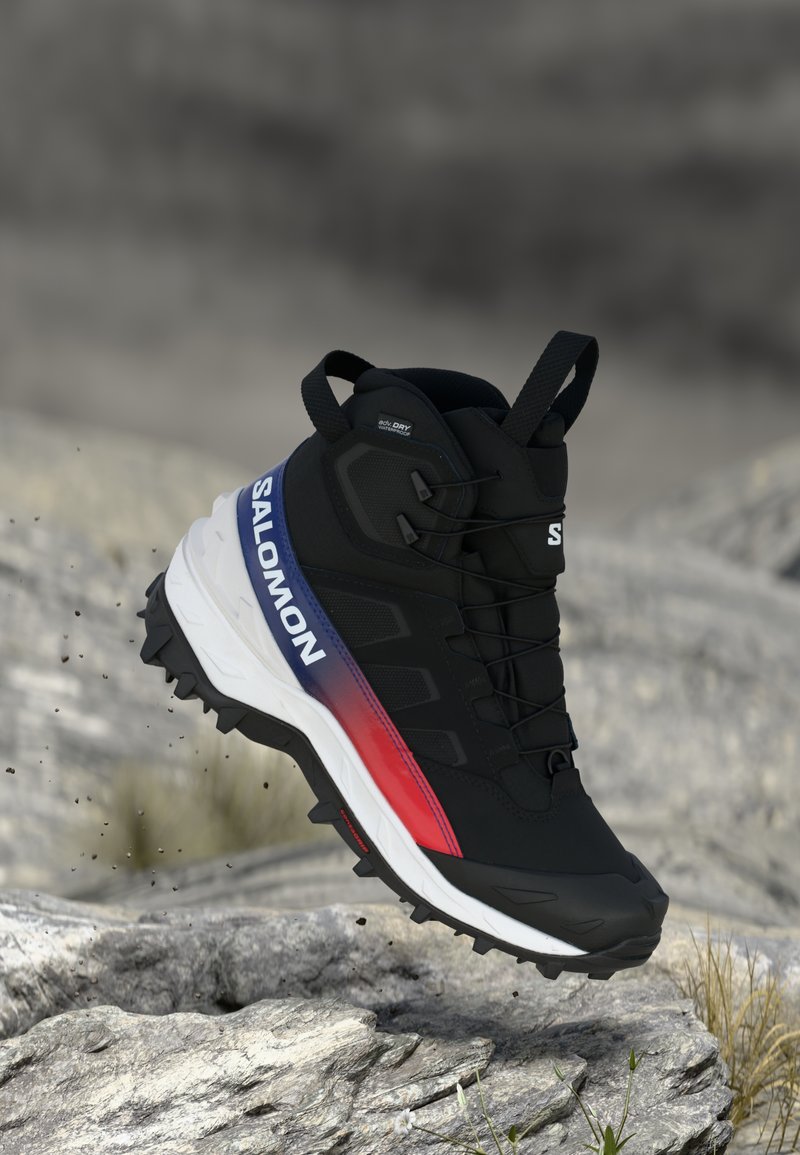Fekete Salomon vízálló túracipő kék és vörös kiegészítőkkel, strapabíró talppal, mid-air (alsó légmentes területen) sziklás talaj felett, apró törmelékek repkednek.