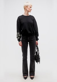 Άτομο με κοντά ξανθά μαλλιά, φοράει μαύρο κεντημένο φούτερ, μαύρο τζιν, μαύρα μυτερά παπούτσια, κρατά μια μαύρη τσάντα, κοιτά προς τα δεξιά.