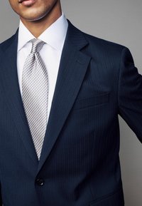 Marineblauer Nadelstreifenanzugjacke mit Reverskragen, getragen über einem weißen Hemd. Eine gemusterte Krawatte in Hellbeige und Blau rundet den Look ab.