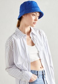 Chapeau seau bleu avec un motif texturé, porté avec une chemise boutonnée à rayures blanches sur un crop top blanc, des shorts en denim, le tout sur un arrière-plan neutre.