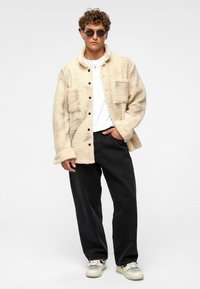 Beige, gestructureerde fleece jas met een kraag en twee voorkanten zakken, gecombineerd met losse zwarte broek en crèmekleurige sneakers.