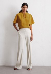 Mustard short-sleeve top with a tie neck, paired with high-waisted, wide-leg cream trousers. Smooth fabric and relaxed fit. Black sandals.